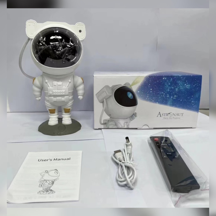عکس از پروژکتور کهکشانی مدل فضانورد همراه با جعبه و کنترل و سیم و دفترچه راهنمای محصول روی میز سفید