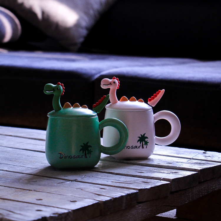 ماگ دایناسوری در دار همراه با قاشق رنگ سبز و صورتی روی میز چوبی