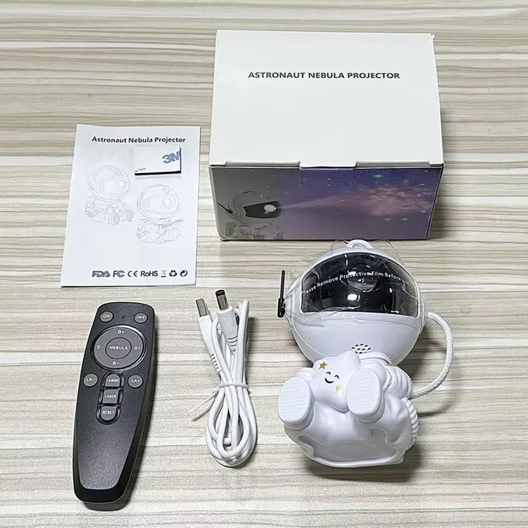 نمایش محتویات اپروژکتور کهکشانی مدل فضانورد نشسته شامل کابل برق - ریموت کنترل - دفترچه راهنما و خود پروژکتور کهکشانی فضانورد نشسته روی میز چوبی