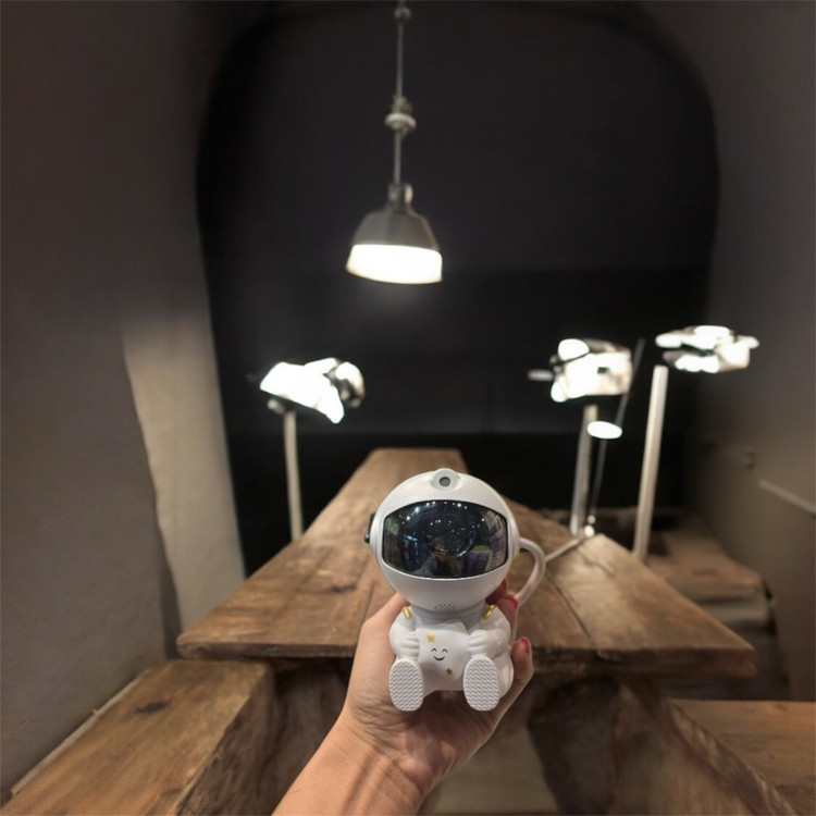 عکس از نمای رو به رو از پروژکتور کهکشانی مدل فضانورد نشسته در دست کاربر با پشت زمینه ی میز چوبی و نور های تصویر برداری